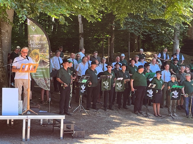 Ein Dirigent dirigiert ein Bläserensemble vor einem Banner mit der Aufschrift "Jagdhornbläser" in einem schattigen Wald. Die Musiker tragen grüne Uniformen und stehen konzentriert aufgestellt.