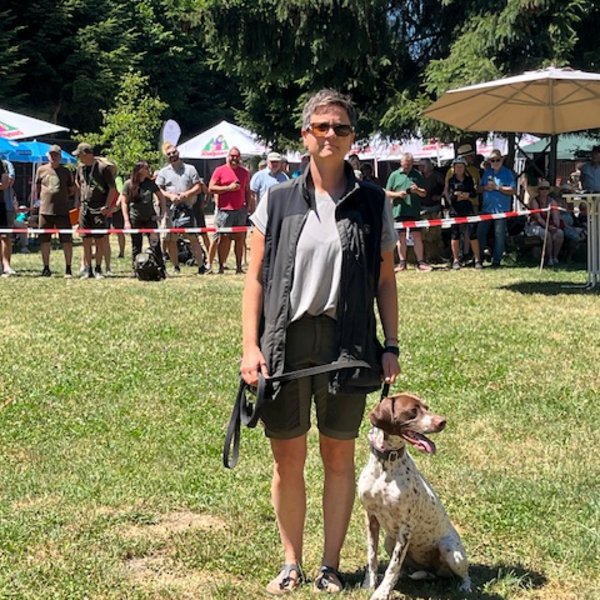 Eine Person hält einen Hund an der Leine. Sie stehen auf einer grünen Wiese, umgeben von Menschen und Ständen bei einem Outdoor-Event im Sonnenschein.