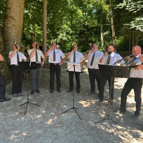 Eine Gruppe Männer spielt Hörner im Freien. Sie tragen weiße Hemden und Krawatten, stehen um Notenständer unter Bäumen auf einem sandigen Untergrund.