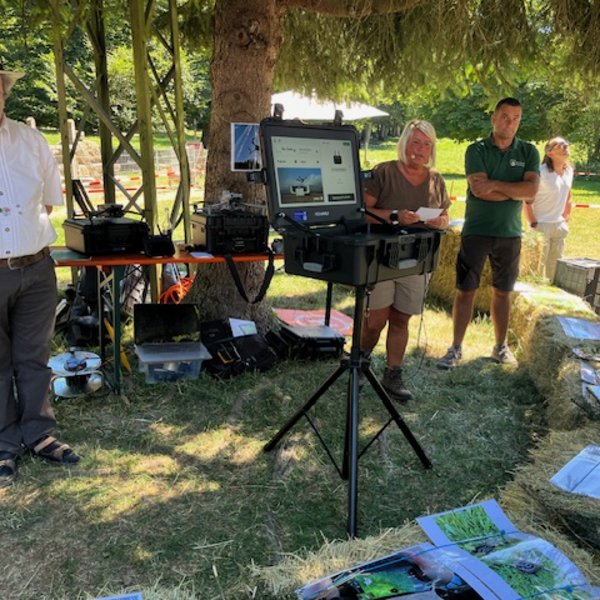 Ein Laptop steht auf einem Stativ, zeigt ein Bild, umgeben von Menschen, Bäumen und Heuballen in einem sonnigen Park. Zahlreiche Geräte und Plakate liegen auf dem Boden.