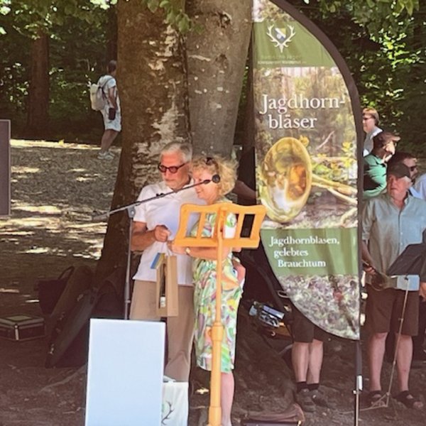 Eine Gruppe von Menschen sitzt an langen, orangefarbenen Bänken im Freien und trägt grüne Hemden. Umgeben von Bäumen, genießen sie das Zusammensein. Ein Banner ist sichtbar, während einige Männer stehen.