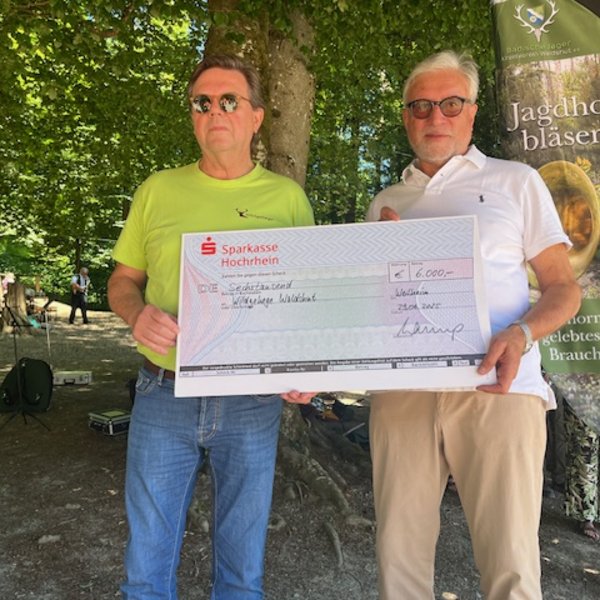Zwei Männer halten einen großen Scheck. Links ist einer in einem grünen T-Shirt, rechts trägt der andere ein weißes Hemd. Der Scheck zeigt: "Sparkasse Hochrhein, 6.000 €, Schutzgemeinschaft Deutscher Wald". Umgebung: Wald mit Infotafel über Jagdhornbläser.