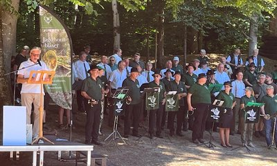 Ein Dirigent dirigiert ein Bläserensemble vor einem Banner mit der Aufschrift "Jagdhornbläser" in einem schattigen Wald. Die Musiker tragen grüne Uniformen und stehen konzentriert aufgestellt.