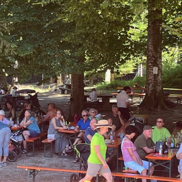 Menschen sitzen auf Bänken in einem schattigen Biergarten unter Bäumen, einige trinken und essen, während andere plaudern. Im Hintergrund sind weitere Besucher und Kinderwagen zu sehen.