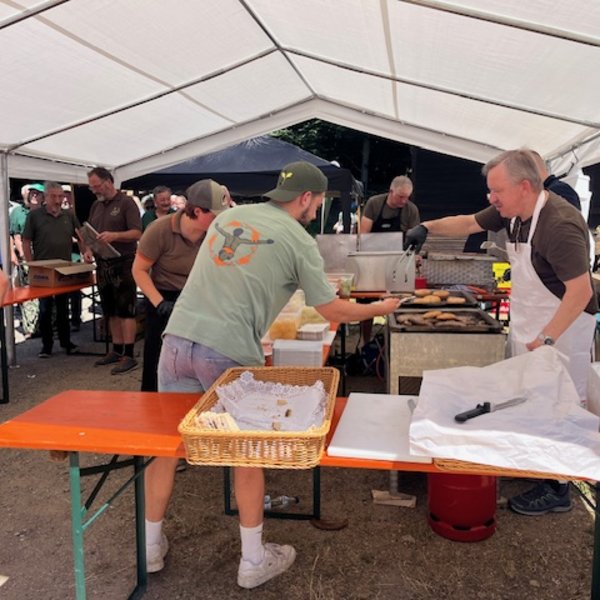 Ein Verkäufer grillt Würstchen unter einem weißen Zelt. Menschen stehen in einer Warteschlange. Ein Tisch mit Brotkorb und Besteck. Im Hintergrund sind Bäume. Text: „GEFAKO“, „Wagner“.