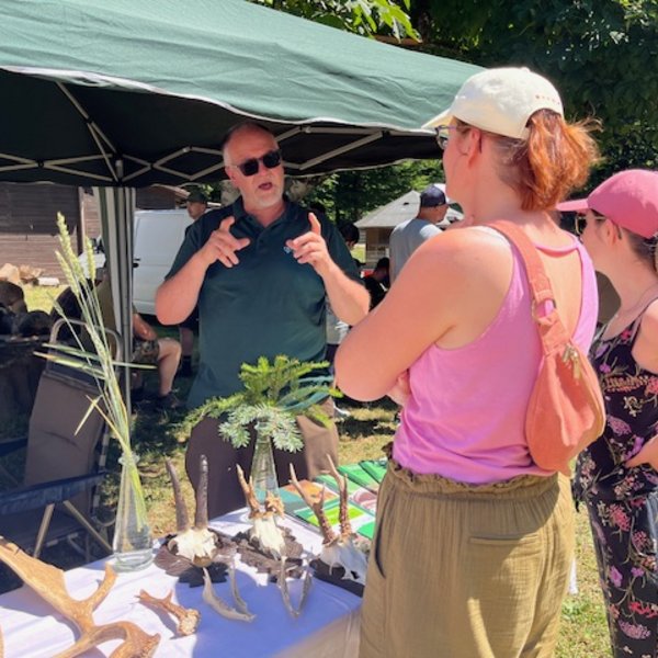 Ein Mann erklärt gestikulierend einigen Frauen Dekorationsgegenstände auf einem Tisch. Sie stehen unter einem grünen Pavillon im Freien bei sonnigem Wetter.