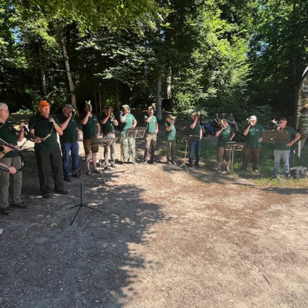 Menschen spielen Blasinstrumente im Freien. Sie stehen in einer Reihe im Wald, umgeben von Bäumen. Ein Banner mit der Aufschrift "Jagdhornbläser" ist sichtbar.